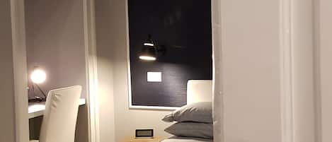 A bedroom with a bed, a desk with a chair, a nightstand, and a window with a view of the night sky.