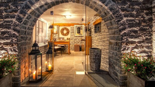 An arched stone doorway leading to a warmly lit interior space with a wooden table, chairs, and a laptop.