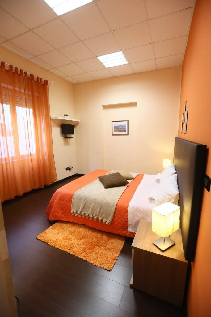 Una habitación de hotel con una cama, una mesita de noche con lámpara, un televisor y una ventana con cortinas.