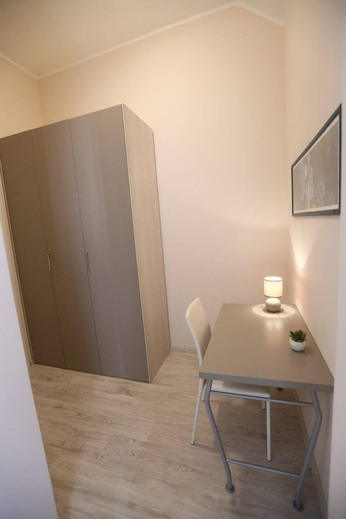 A small room with a single table, a chair, a lamp, a wardrobe, and a framed picture on the wall.