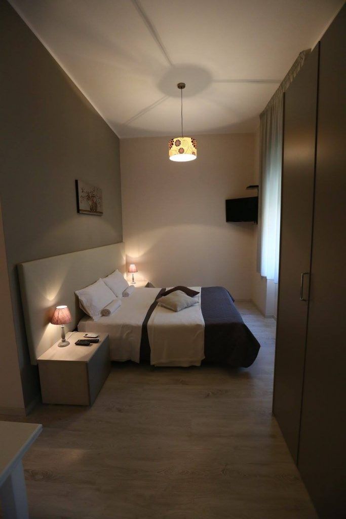 A small, well-lit bedroom with a single bed, bedside tables, a hanging light fixture, and a television mounted on the wall.
