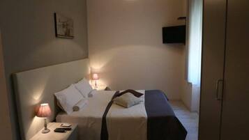 A small, well-lit bedroom with a single bed, bedside tables, a hanging light fixture, and a television mounted on the wall.