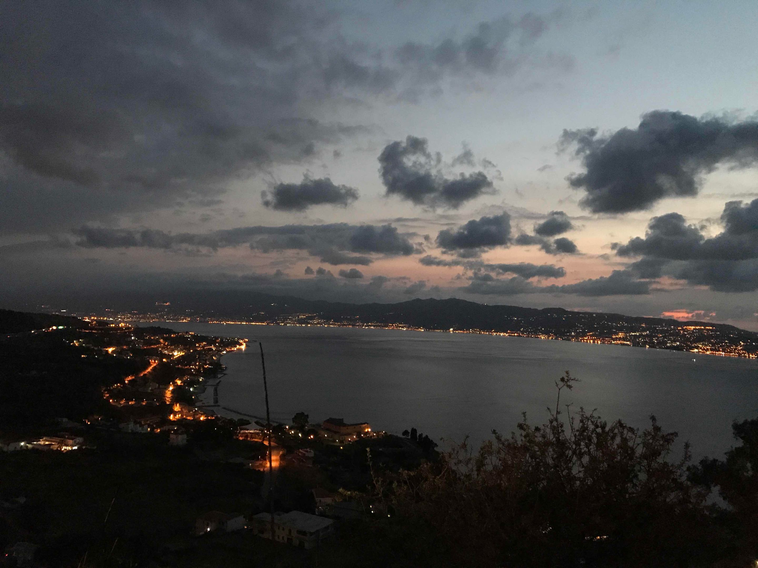 A coastal cityscape at dusk with a view of the sea and distant mountains.