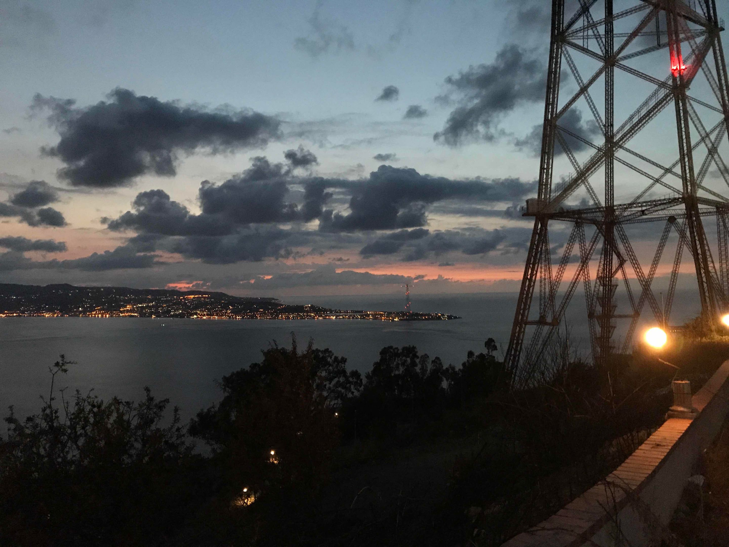 A coastal cityscape at dusk with a prominent electricity pylon and distant city lights.