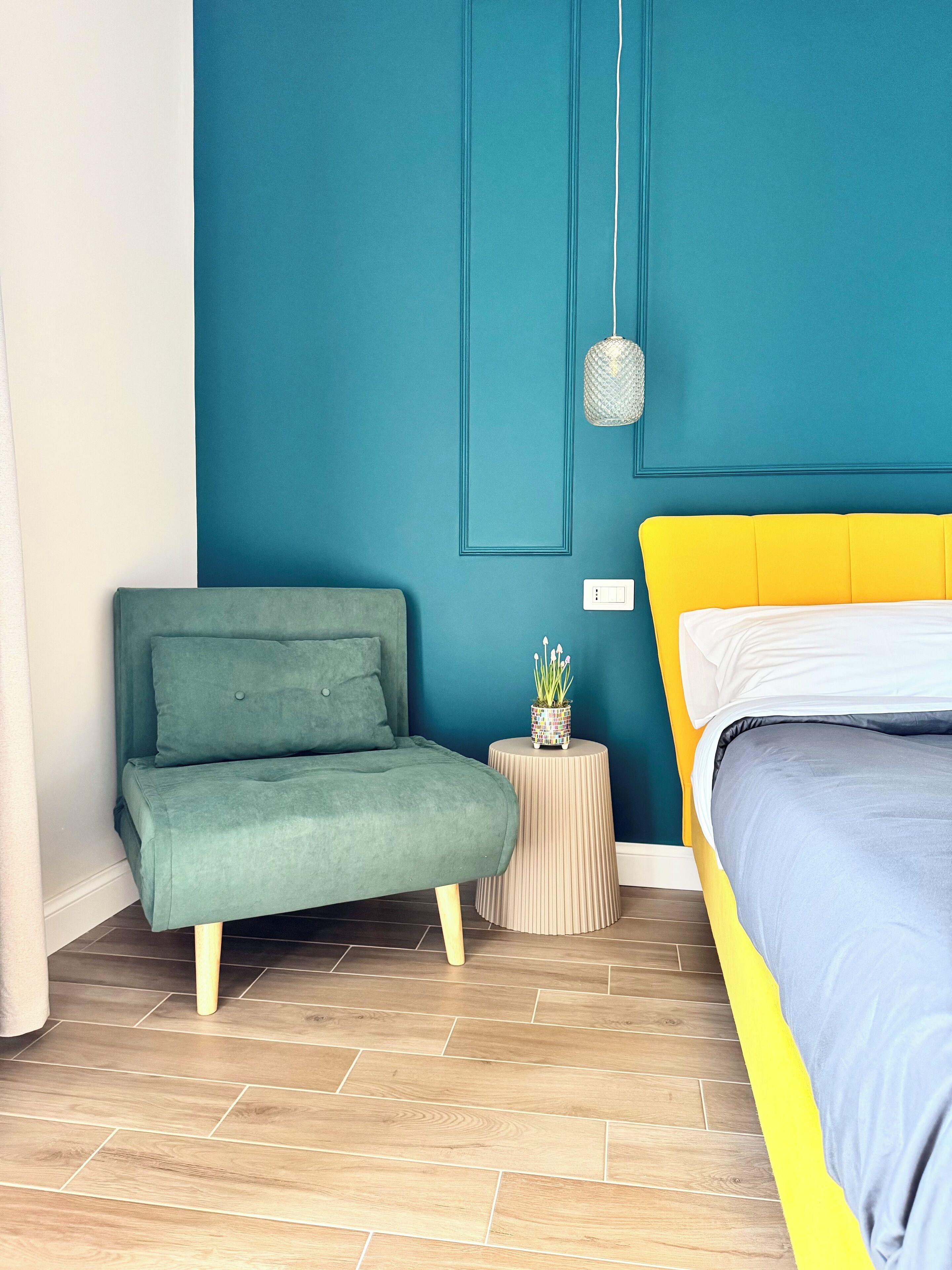 Un dormitorio con una pared de acento color aguamarina, un sofá verde, una cama amarilla y una pequeña mesa redonda con una planta.