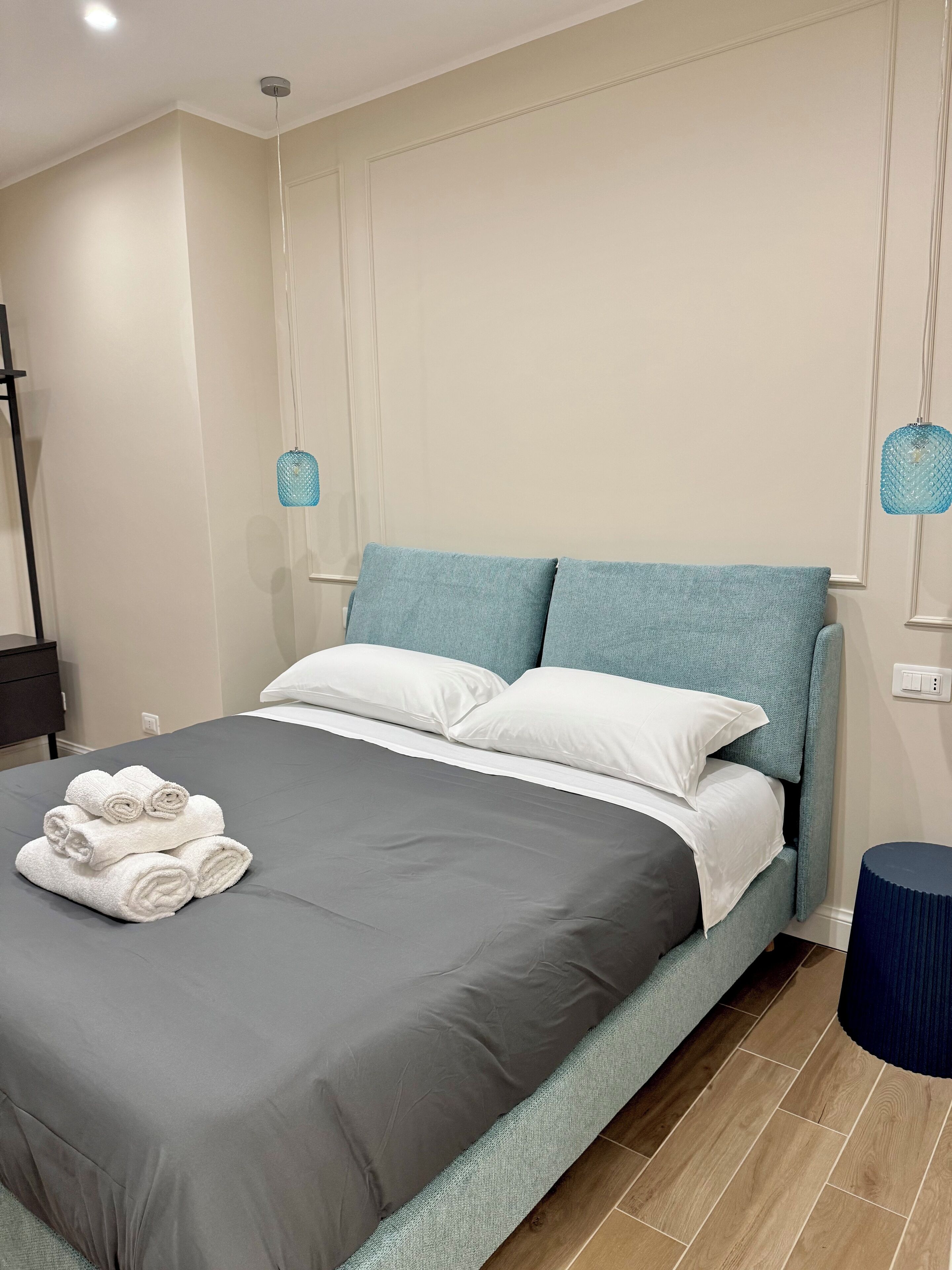 A neatly made bed with a grey bedspread and white pillows, a blue headboard, and a blue lamp on either side.
