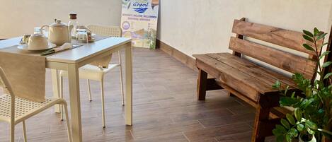 A dining area with a table set for two, a wooden bench, and a potted plant. A sign with multilingual welcome messages is visible in the background.