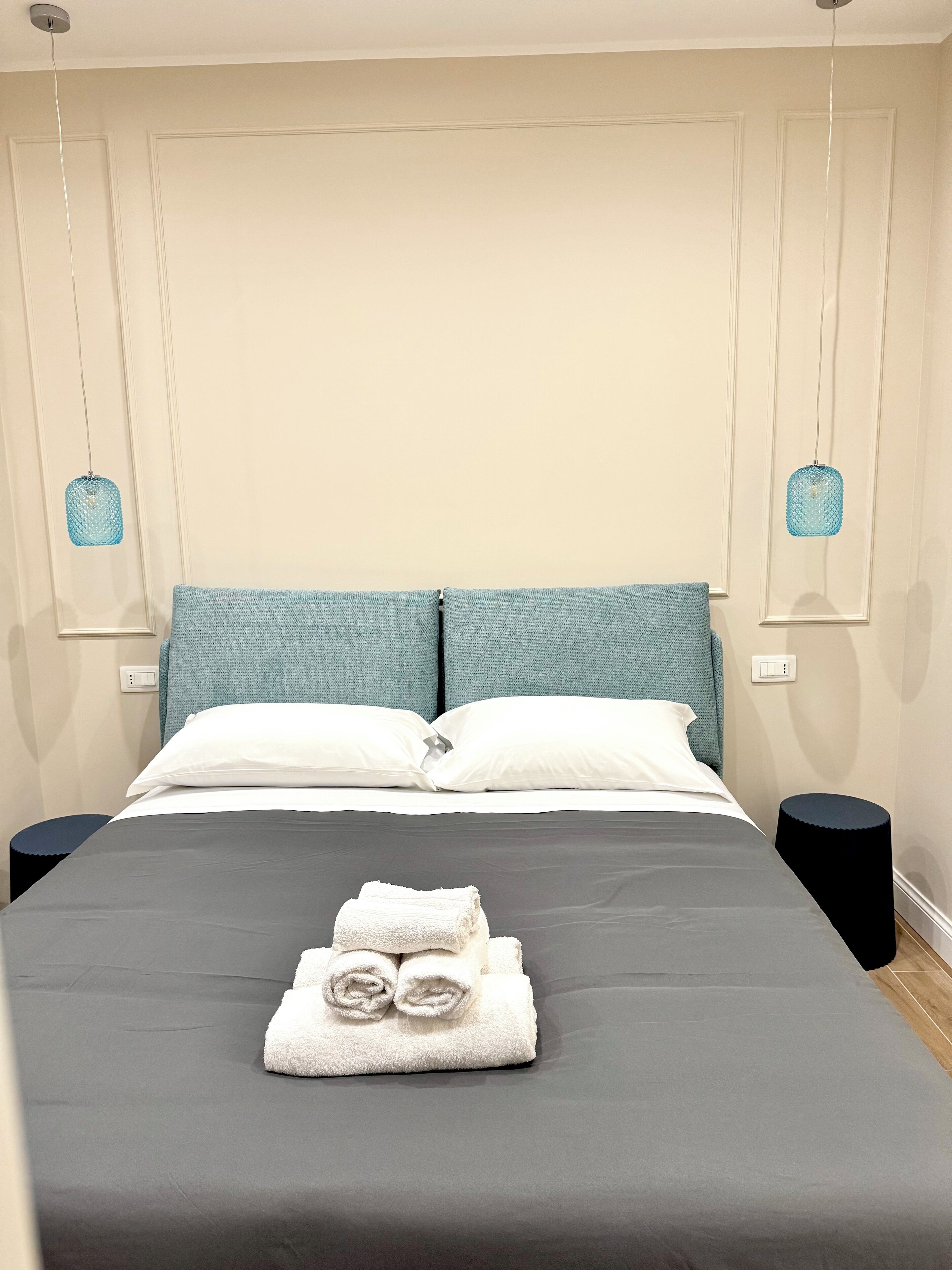 Una cama bien hecha con una colcha gris, dos almohadas blancas y una toalla blanca doblada sobre la cama. Hay dos mesitas de noche negras con una lámpara azul en cada una. La habitación tiene paredes color crema y un diseño sencillo y limpio.