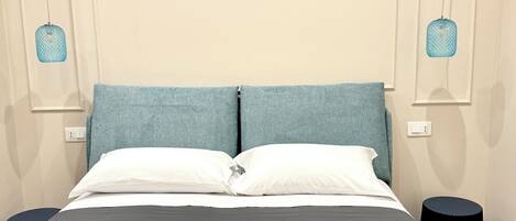 Una cama bien hecha con una colcha gris, dos almohadas blancas y una toalla blanca doblada sobre la cama. Hay dos mesitas de noche negras con una lámpara azul en cada una. La habitación tiene paredes color crema y un diseño sencillo y limpio.