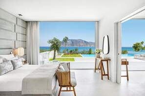 A modern bedroom with a large bed, a wicker chair, a small table, and a view of the sea and mountains.