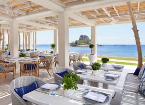 An outdoor dining area with tables and chairs, a view of the sea, and a rocky island in the distance.