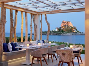 Un área de comedor al aire libre con mesas y sillas, vista al mar y una isla rocosa distante.
