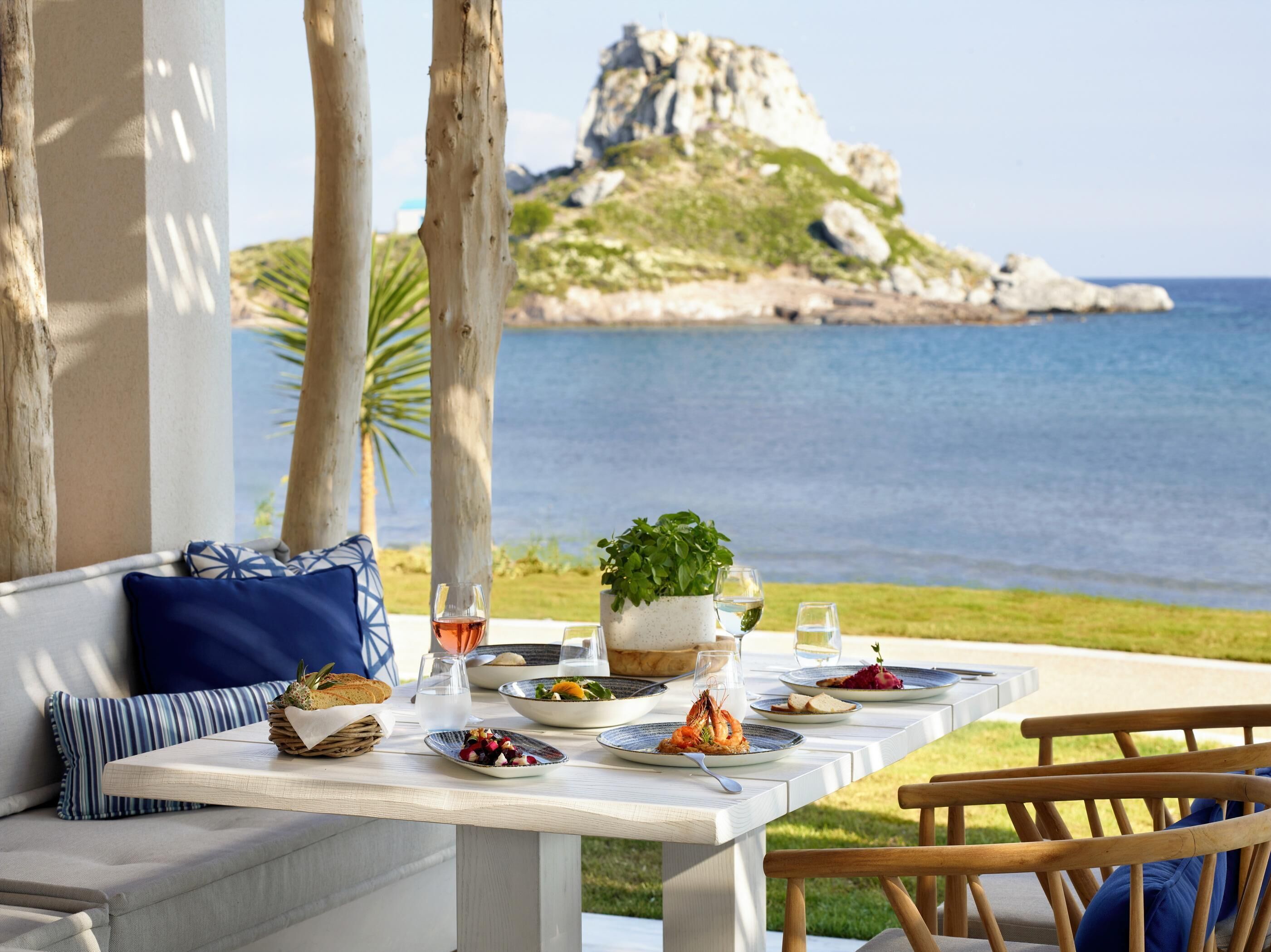 A dining table set with plates of food, glasses, and a potted plant, overlooking a body of water and a rocky landscape.