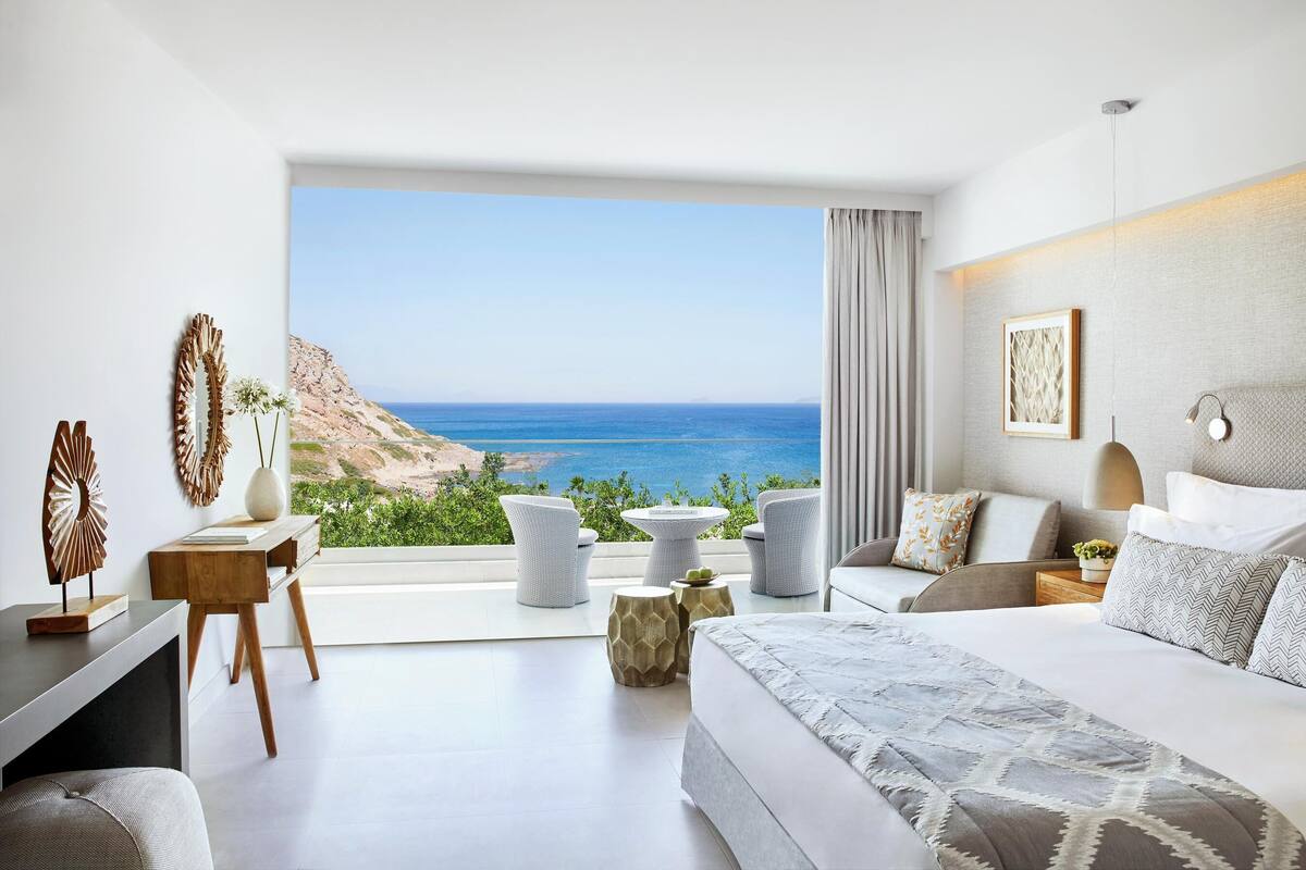 A modern bedroom with a large window overlooking a coastal view, a bed, a desk, and a seating area with chairs.