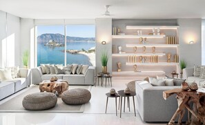 A modern living room with a large window overlooking a coastal view, featuring sofas, ottomans, and a wooden coffee table. - Ikos Aria - All Inclusive (Kos)