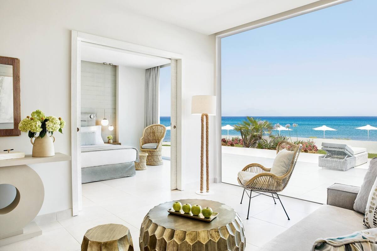 A modern hotel room with a balcony overlooking the sea, a bed, a sitting area with wicker furniture, and a dining table with apples.