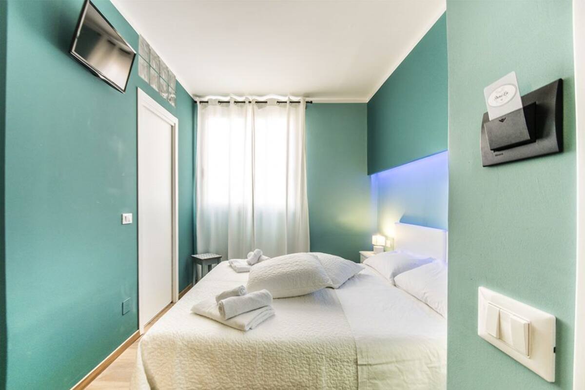 Ein Hotelzimmer mit Türkiswänden, einem Bett mit weißer Bettwäsche, einem an der Wand befestigten Flachbildfernseher, einem Fenster mit weißen Vorhängen und einem Nachttisch mit Lampe.