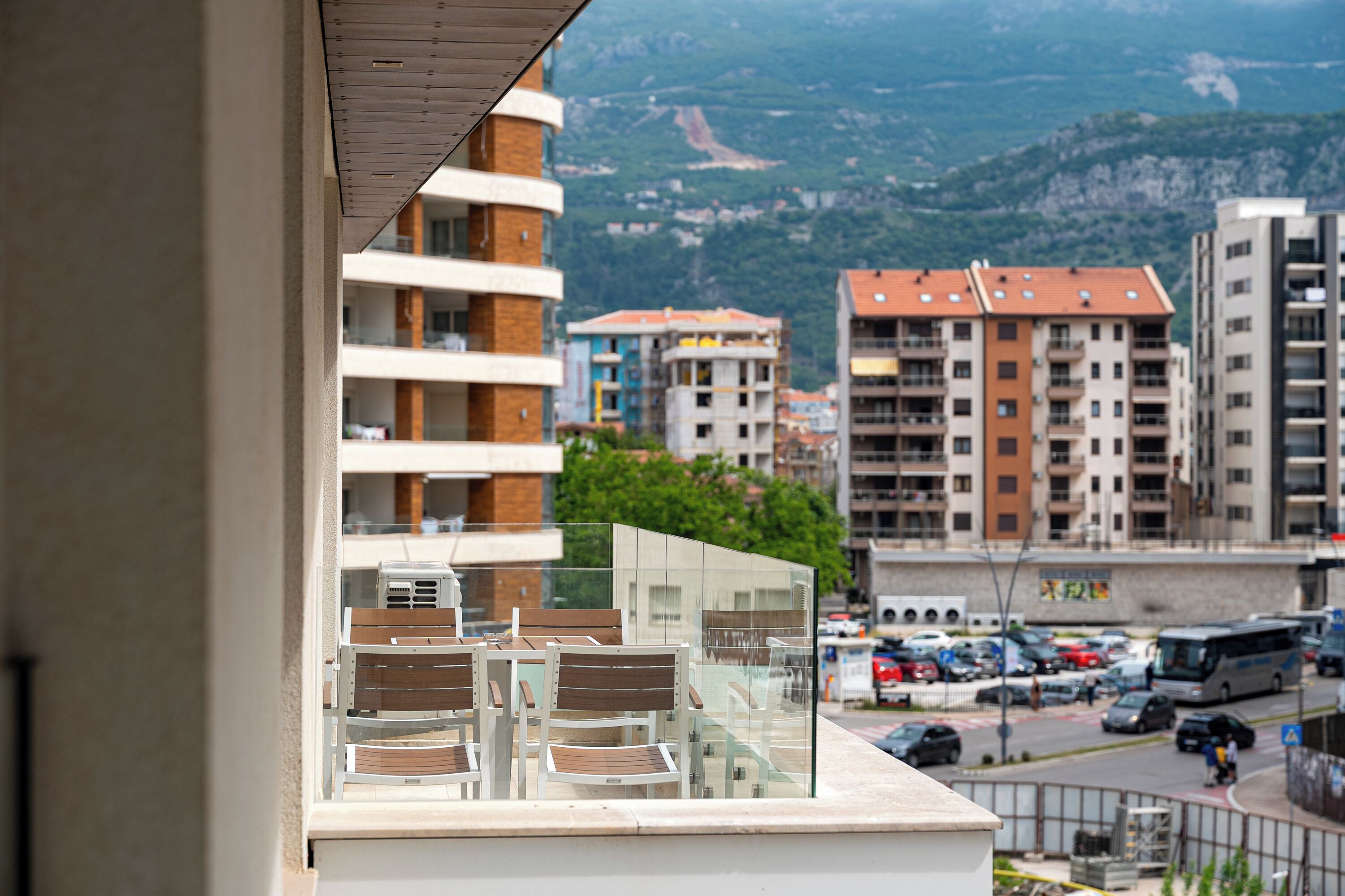 Un balcó amb cadires i una partició de vidre, amb vista a un carrer amb edificis i un paisatge de muntanya.