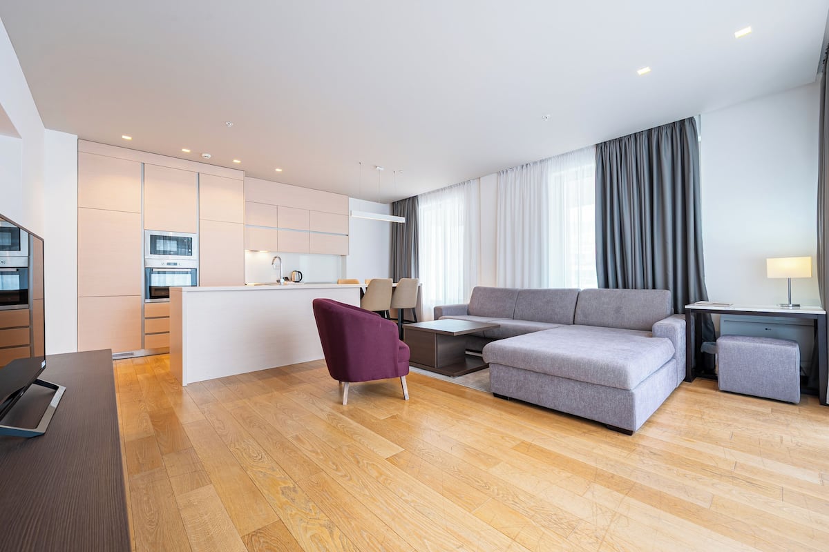 A modern living room with a grey sofa, a purple armchair, a wooden floor, a kitchen area, and a large window with curtains.