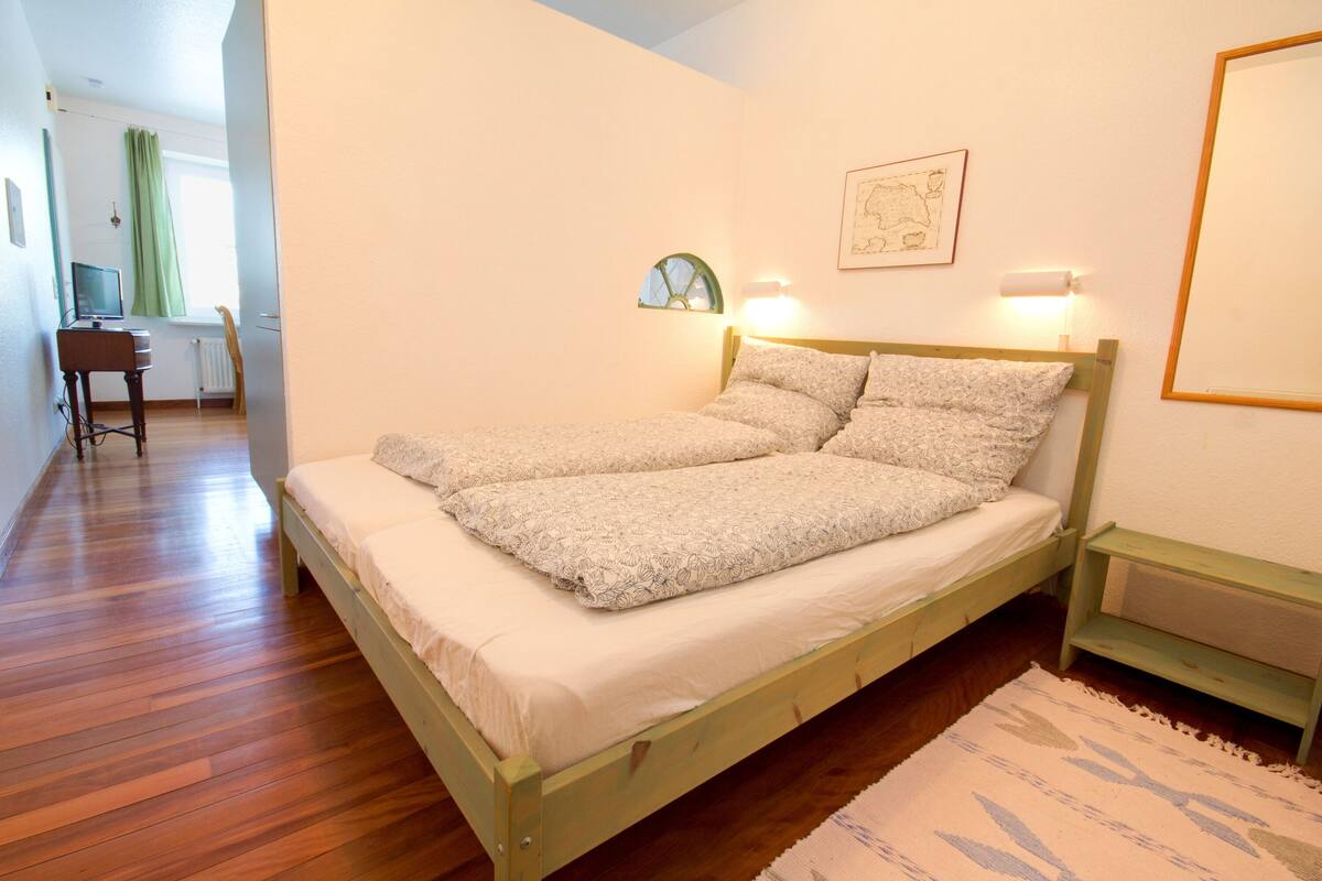 A single bed with a wooden frame, a bedside table, a wall-mounted lamp, and a small window with a curtain.