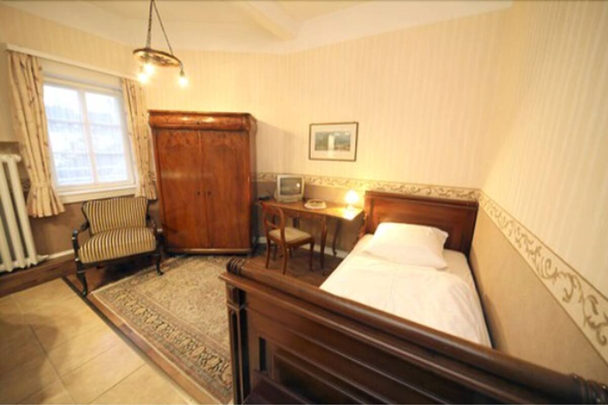 A bedroom with a wooden bed, a chair, a small table with a lamp, a radiator, and a window with curtains.