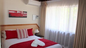 A hotel room with a bed, red and white bedding, a red throw blanket, a folded towel, a nightstand with a lamp, a wall-mounted air conditioner, a framed picture, and sheer curtains on a window. - Flamingo Lodge (St. Lucia)