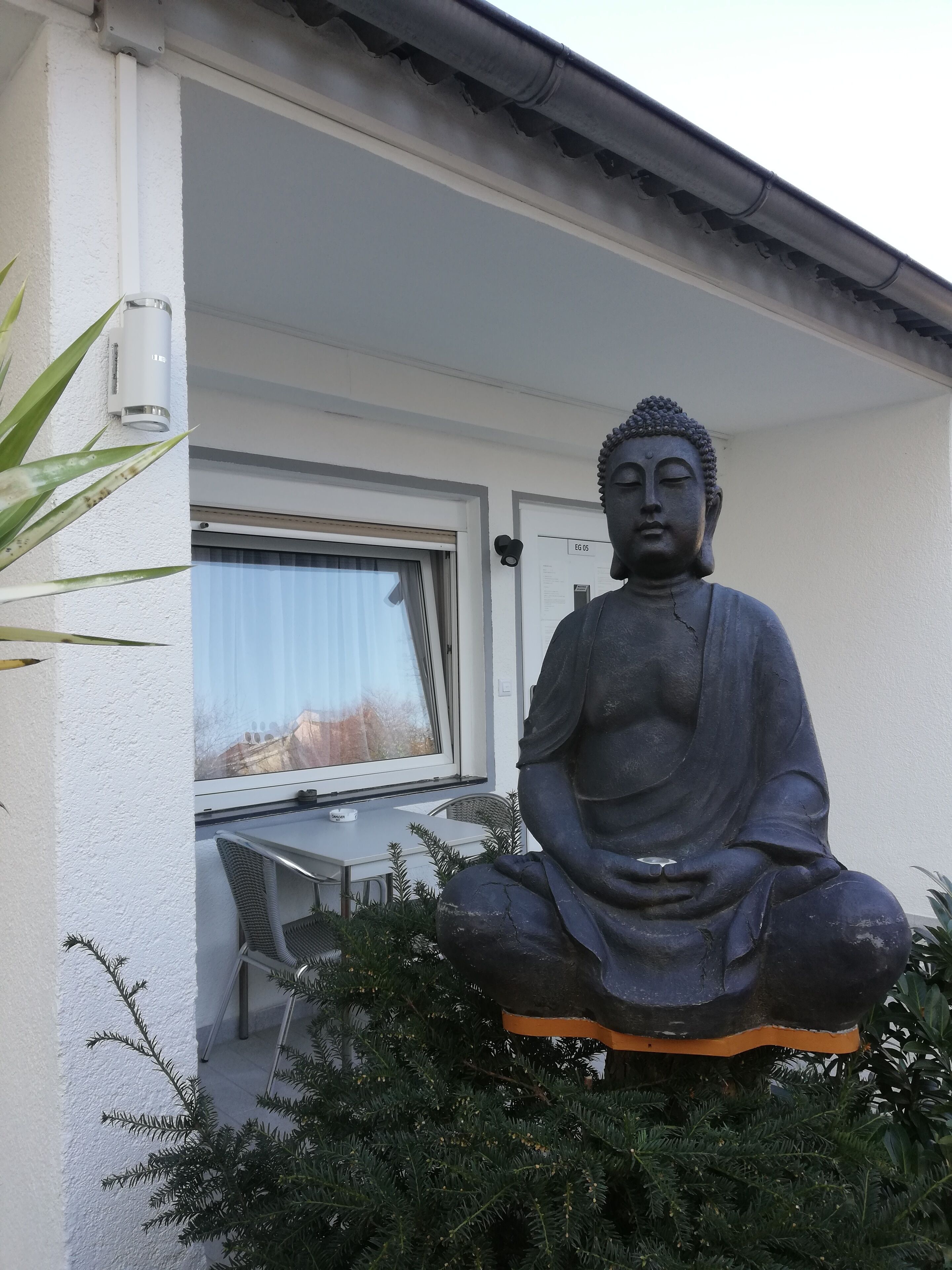 A Buddha statue in a garden area with a table and chairs nearby, a window with a view, and a white wall.