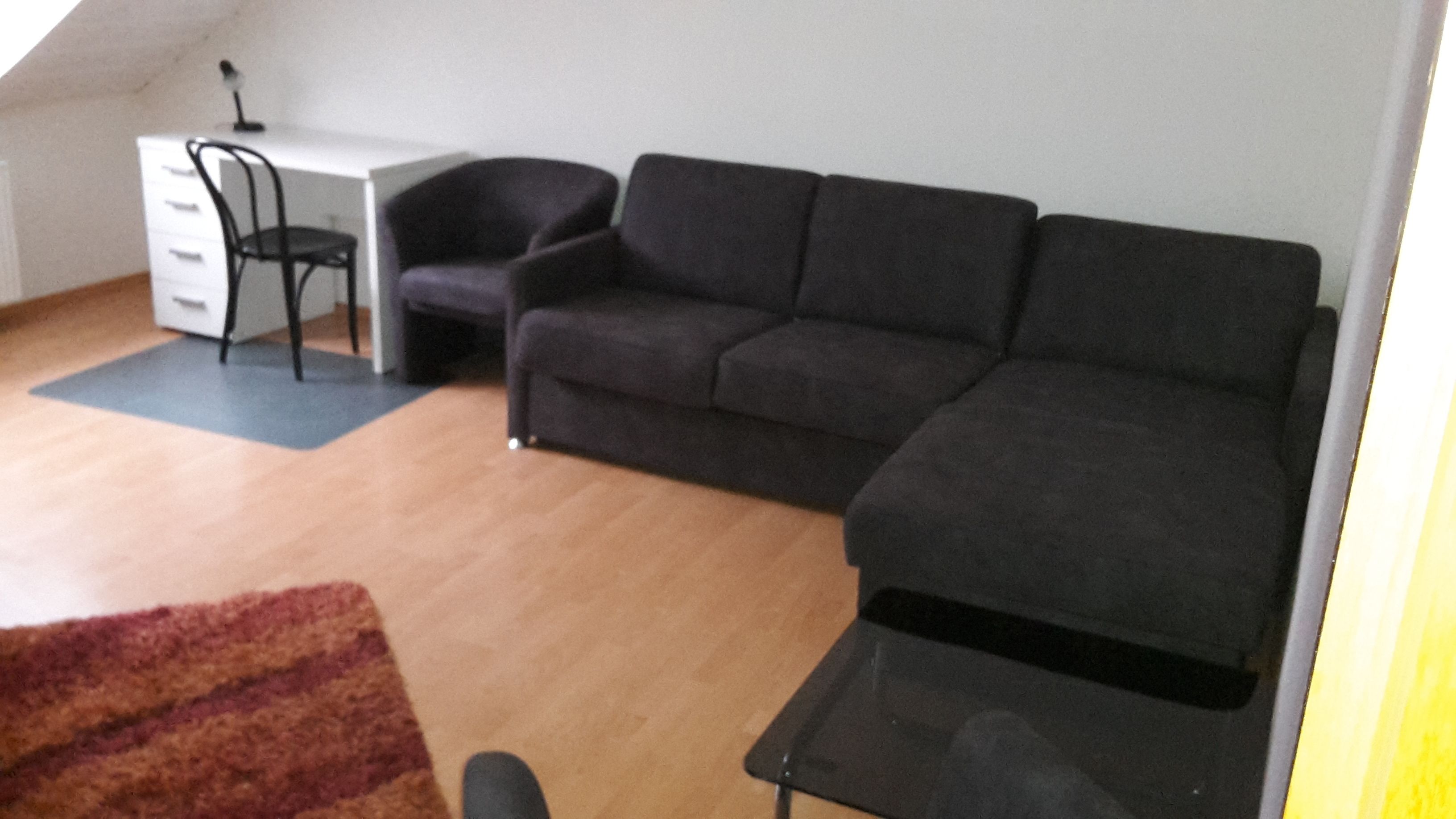 Una sala de estar con un sofá negro, un escritorio blanco, una silla negra y piso de madera.