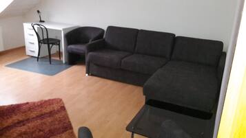 Una sala de estar con un sofá negro, un escritorio blanco, una silla negra y piso de madera.