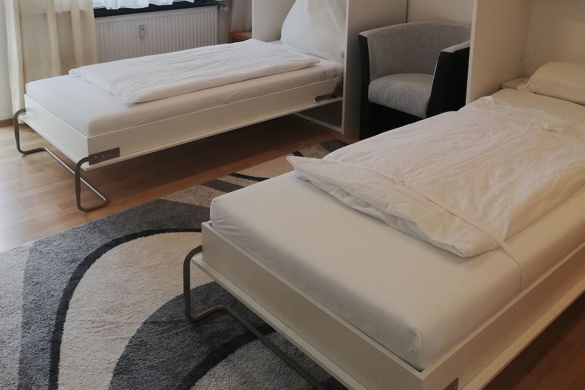 Habitación de hotel con dos camas individuales, un sillón gris, un armario blanco y una ventana con cortinas traslúcidas.