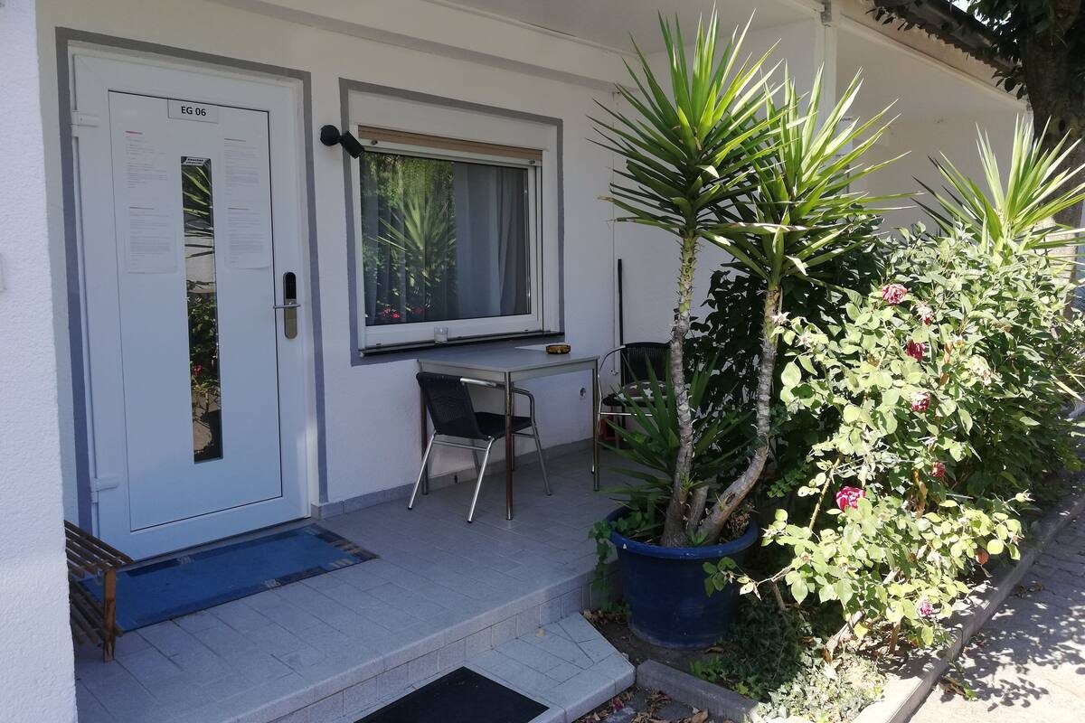 Un pequeño edificio de una sola planta con fachada blanca, una puerta con la leyenda “EG 06”, una ventana, una mesa pequeña con una silla y una planta en maceta con hojas verdes y flores rojas.