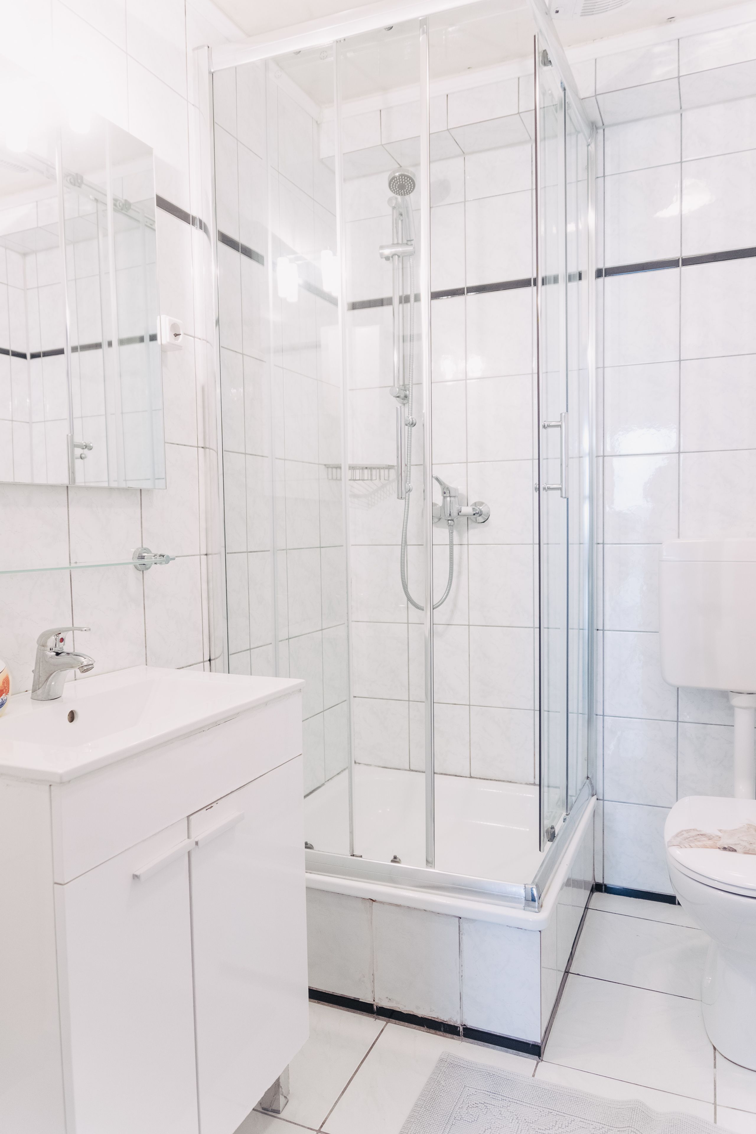 Un baño moderno con ducha de vidrio, azulejos blancos, un lavamanos blanco y un inodoro.