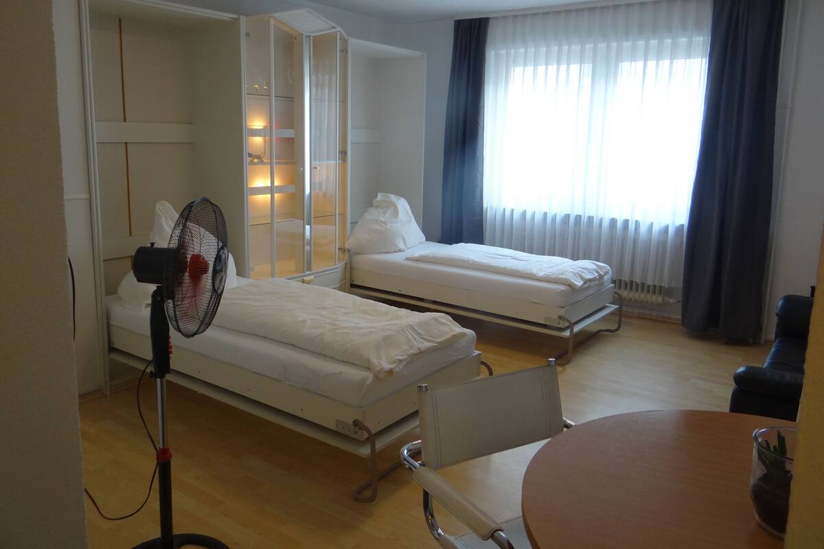 Habitación de hotel con dos camas, una mesita de noche, una silla, un ventilador y una ventana con cortinas.