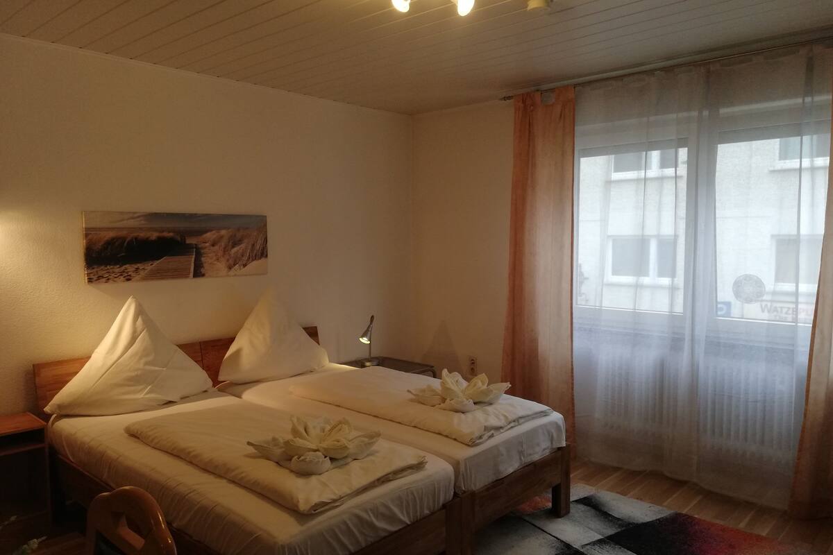 Habitación de hotel con dos camas, una mesita de noche, una lámpara, un cuadro en la pared y una ventana con cortinas.