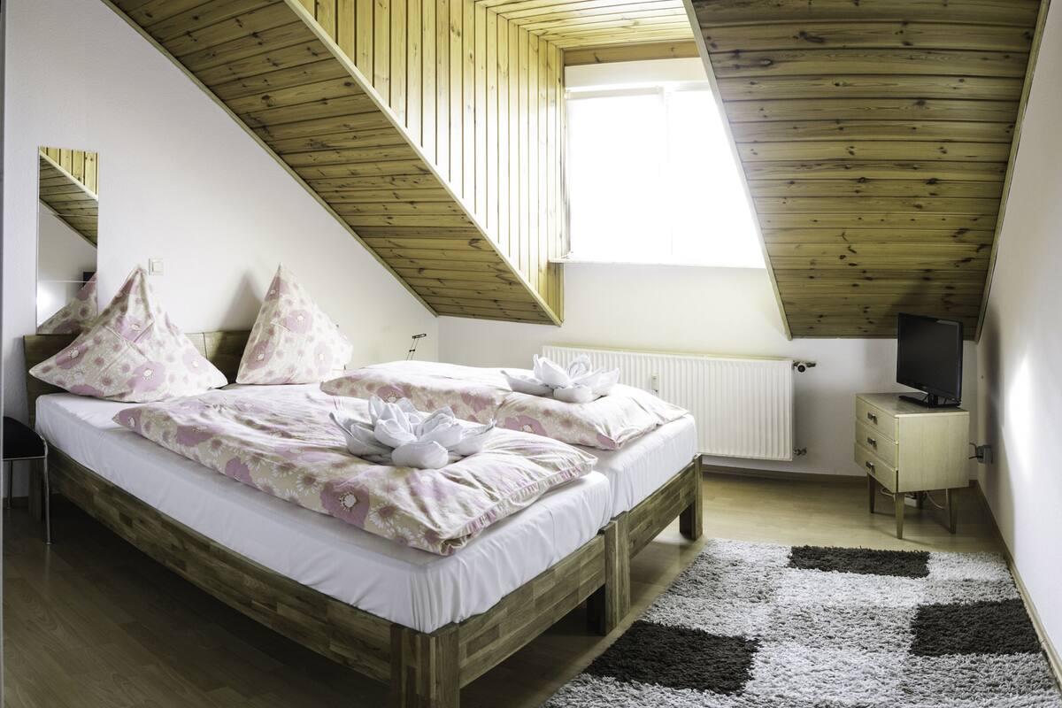 Un dormitorio con techo inclinado de madera, una cama con ropa de cama floral, una mesita de noche, un radiador y un televisor sobre un aparador.