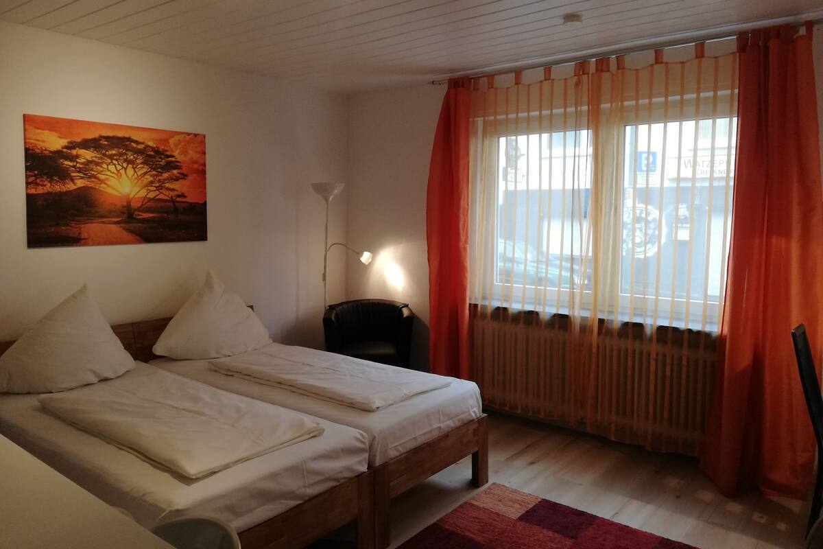 Una habitación con dos camas, una lámpara de pie, un cuadro de un atardecer y una ventana con cortinas.