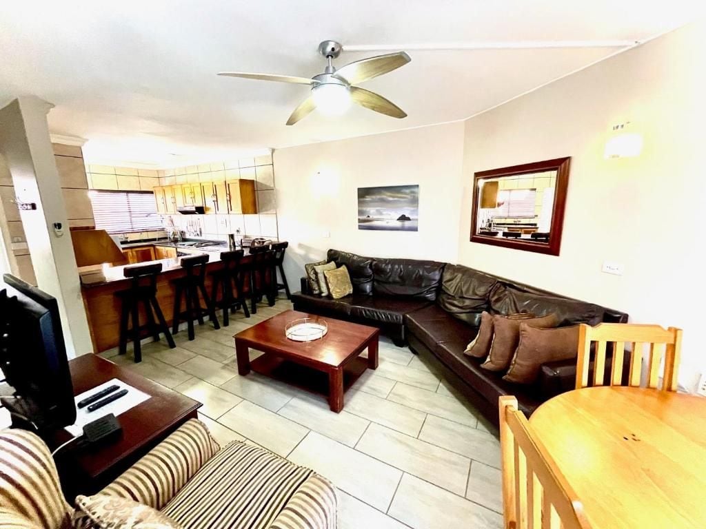 Een woonkamer met een bank, salontafel en een televisie. Er is een plafondventilator, een keuken op de achtergrond en een eethoek met stoelen.