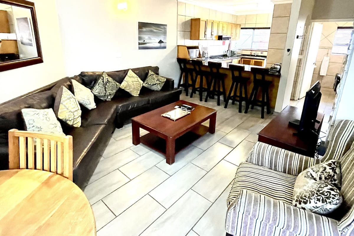 A living room with a sofa, a coffee table, and a striped armchair. There is a kitchen area with a bar and a ceiling fan.
