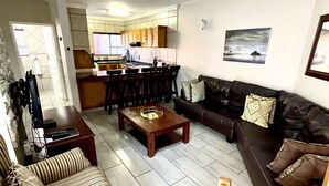 A living room with a sofa, armchair, coffee table, and a ceiling fan. There is a kitchen in the background with a dining area.