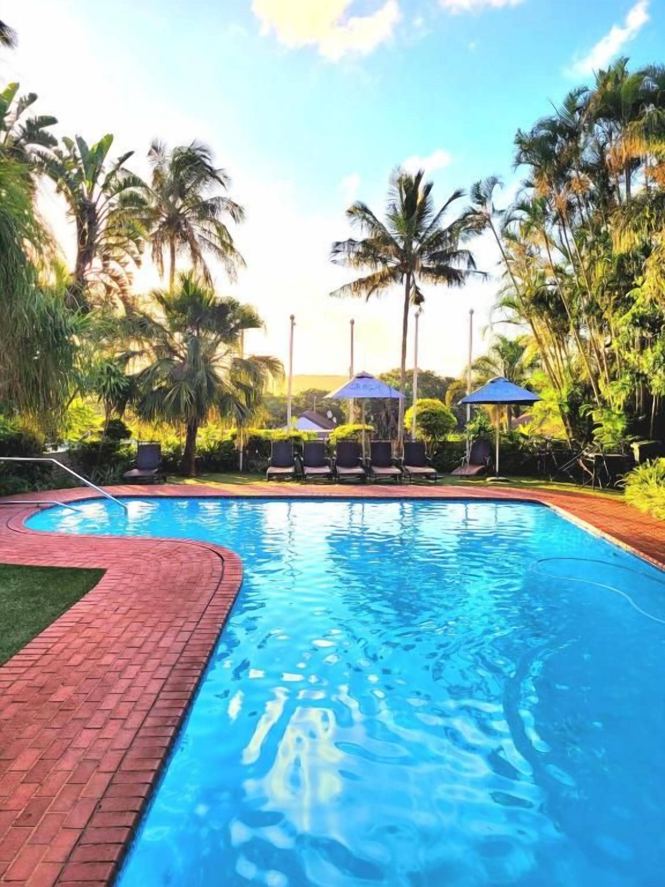 Una piscina con borde de ladrillo, rodeada de palmeras y sillas de descanso.