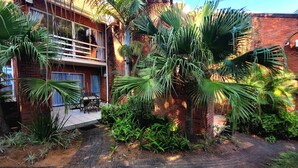 A brick building with a balcony, surrounded by lush greenery including palm trees and various plants.