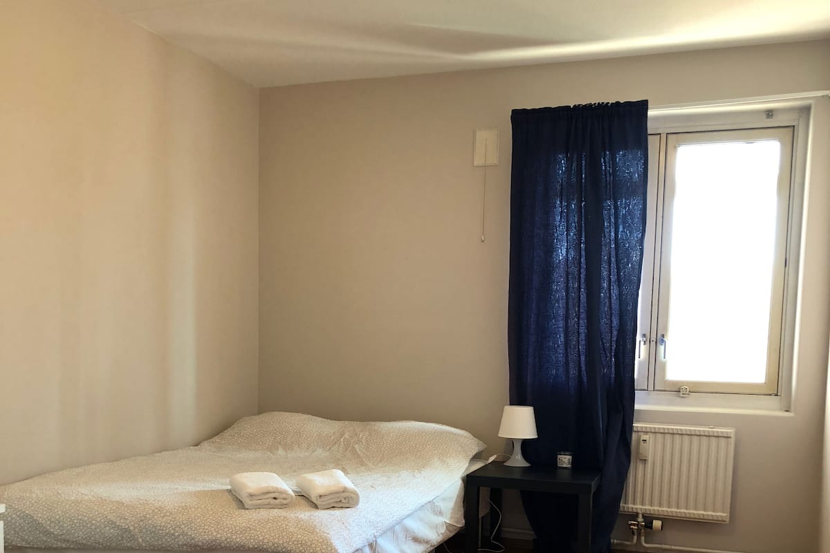 A small, tidy room with a single bed, a nightstand, a lamp, and a window with curtains.