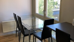 A room with a glass-top dining table, black chairs, a radiator, and a large window with a view of buildings and trees. - Guest House Centrum 3 (Oslo)
