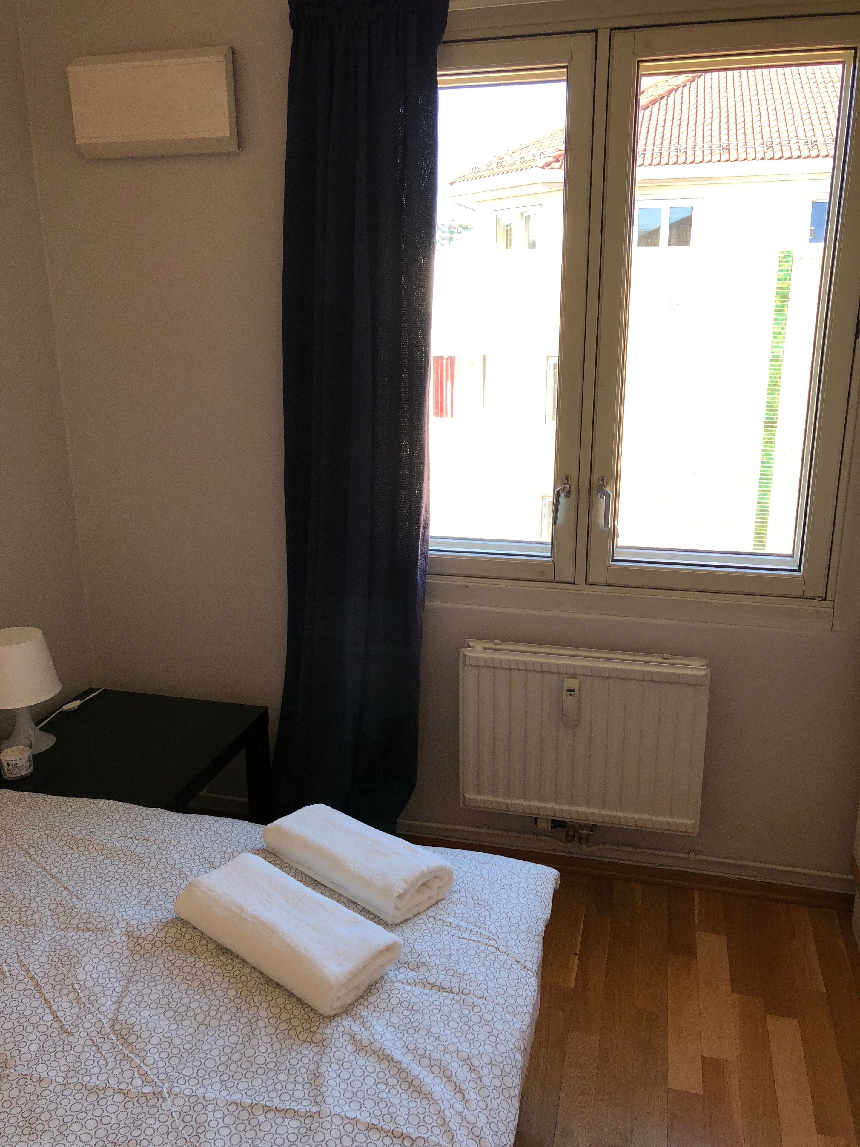 A bedroom with a bed, a nightstand, a window with curtains, and a radiator.