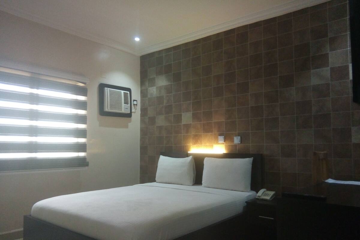 A hotel room with a bed, a nightstand, a wall-mounted air conditioning unit, and a window with blinds.