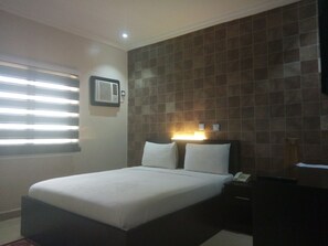 Een hotelkamer met een bed, nachtkastje, wandairconditioning en een raam met zonwering.