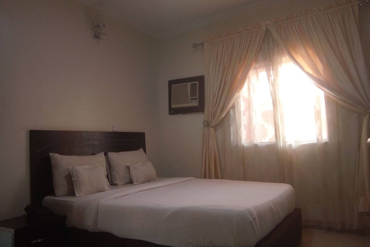 A bedroom with a single bed, a nightstand, a wall-mounted air conditioning unit, and a window with curtains.