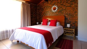A hotel room with a brick wall, a wooden ceiling, a bed with white and red bedding, a red throw blanket, a wooden nightstand with a lamp, and a red rug. - St Lucia Palms (St. Lucia)