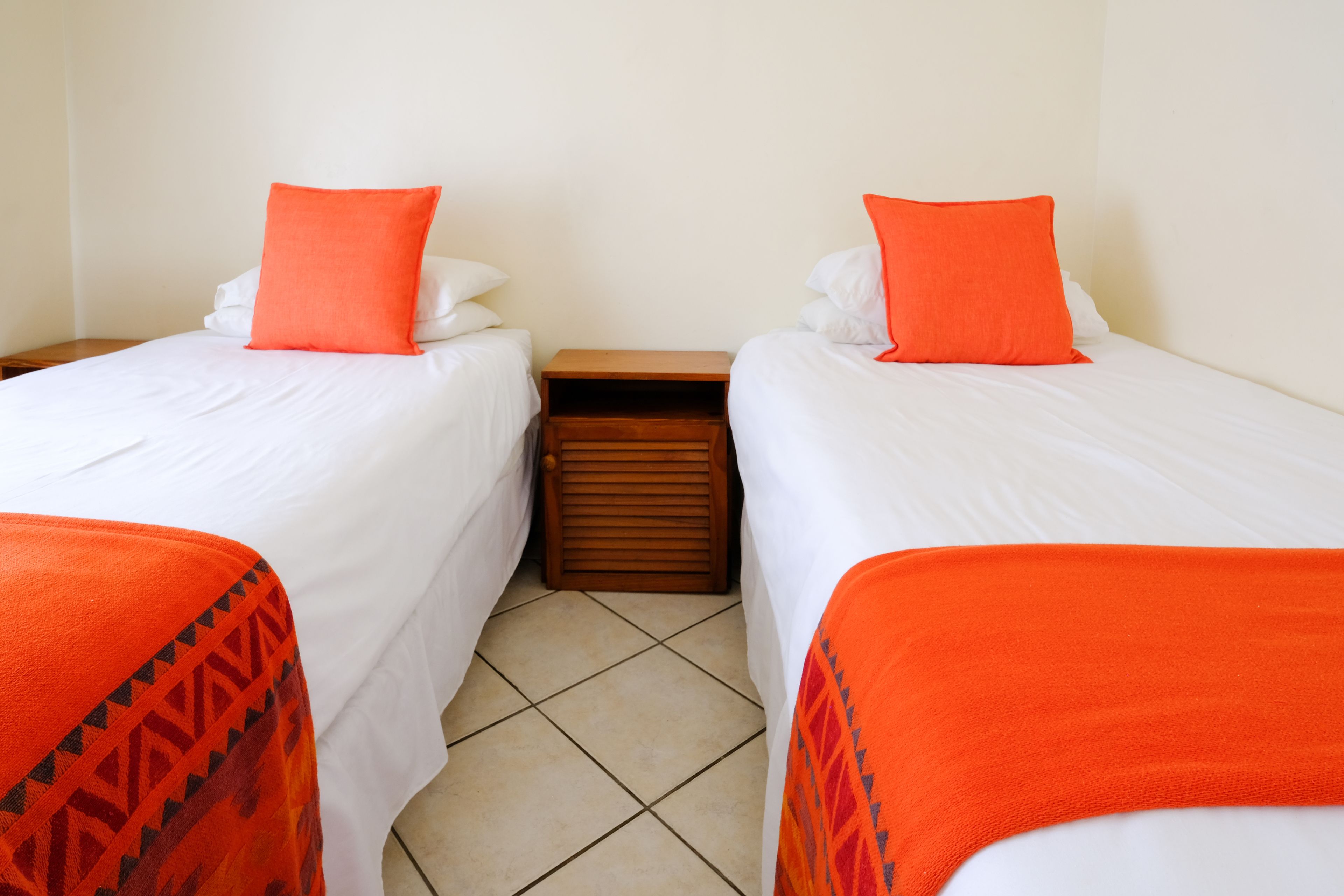 Quarto com duas camas, cada uma com roupa de cama branca e almofadas laranja. Um criado-mudo de madeira fica entre as camas.