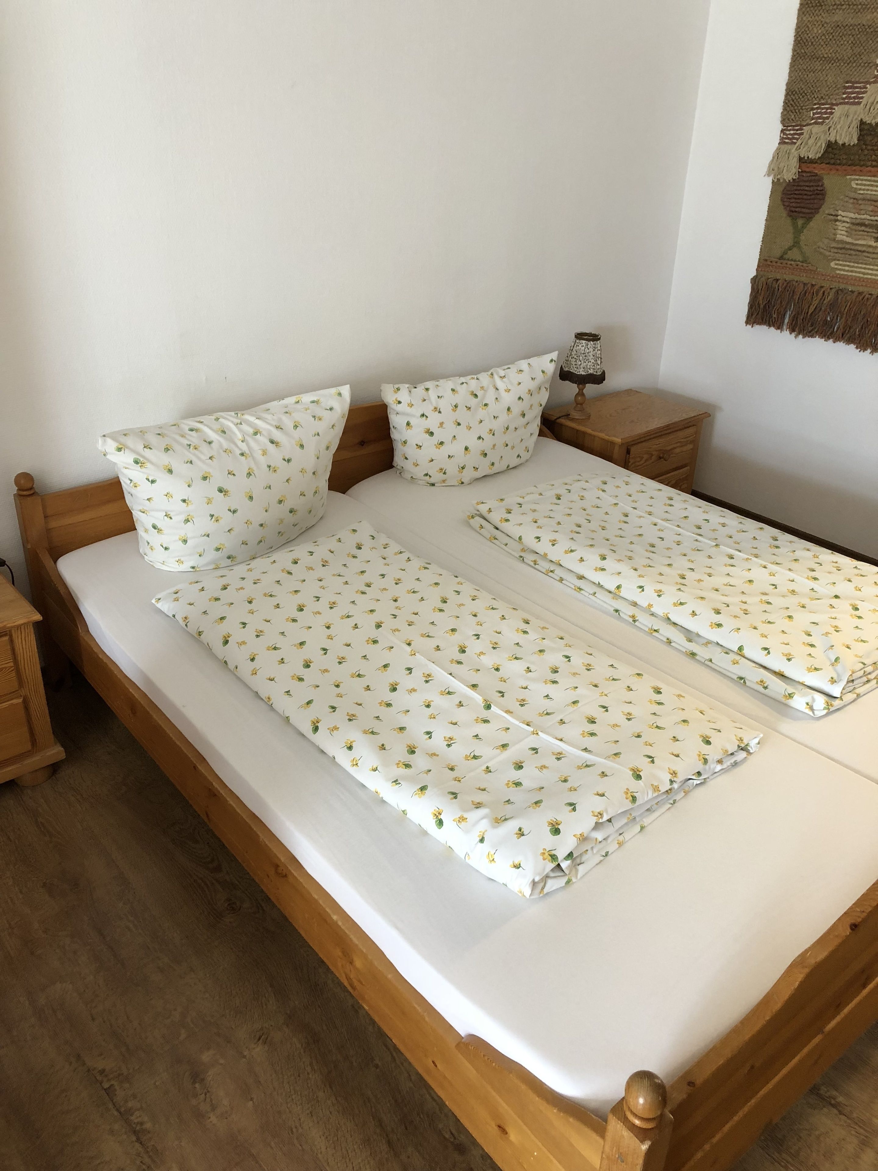 A double bed with a floral-patterned bedspread and pillows, wooden headboard, two bedside tables with lamps, and a woven tapestry on the wall.